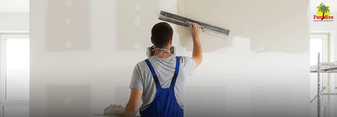 A construction worker wearing a respirator and blue overalls is seen from the back, applying plaster or joint compound to a large white interior wall with a wide metal trowel.