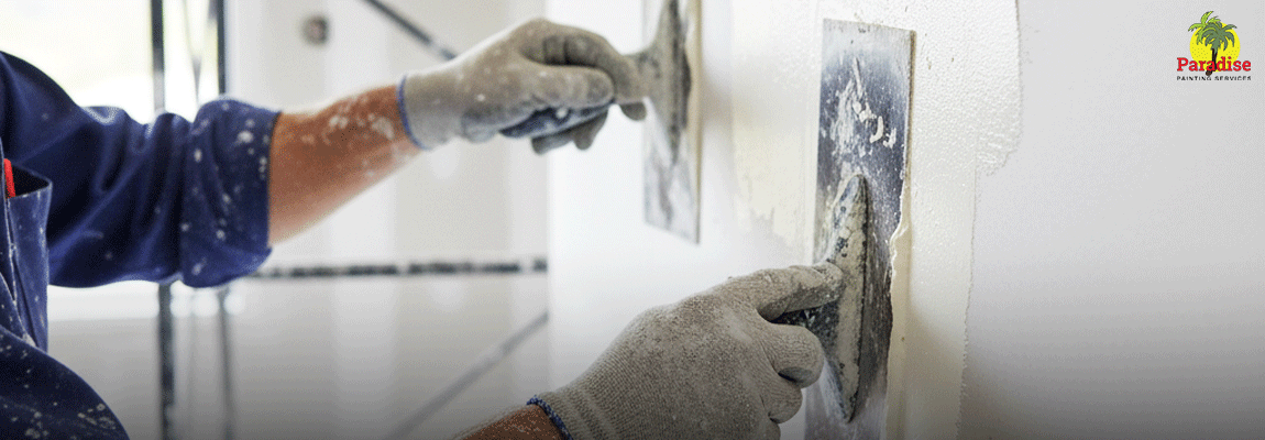Close-up of a worker's hands in gray gloves applying a layer of wet plaster or joint compound to a white wall using two metal plastering trowels.